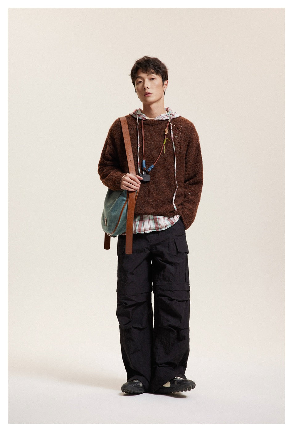 Model facing forward in the brown distressed knit sweater layered over plaid hooded top with black cargo pants