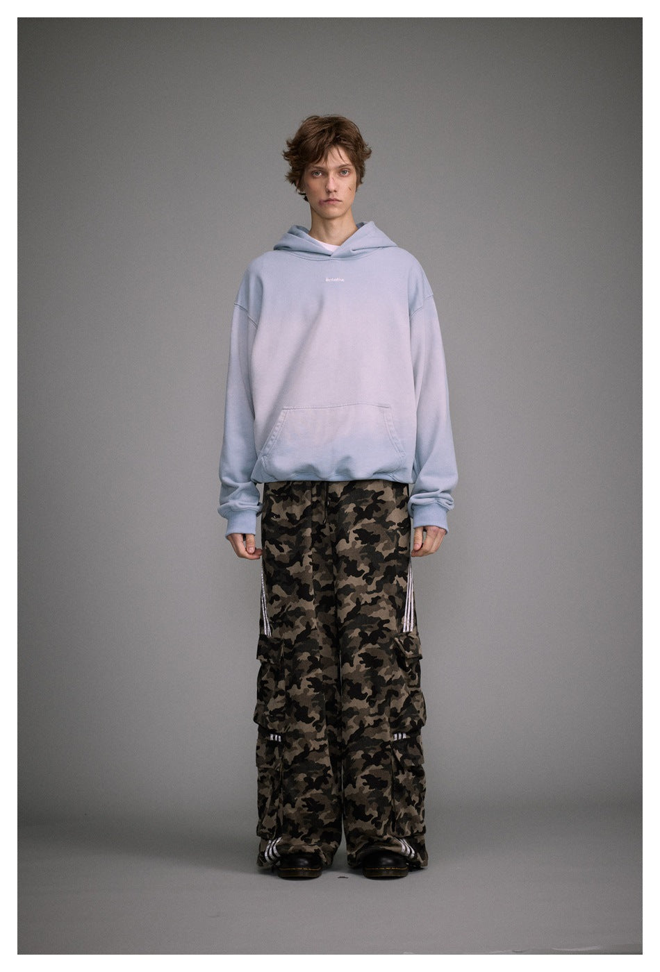 Model wearing an oversized pale blue hoodie with small white “breathe” embroidery and loose camouflage cargo pants.