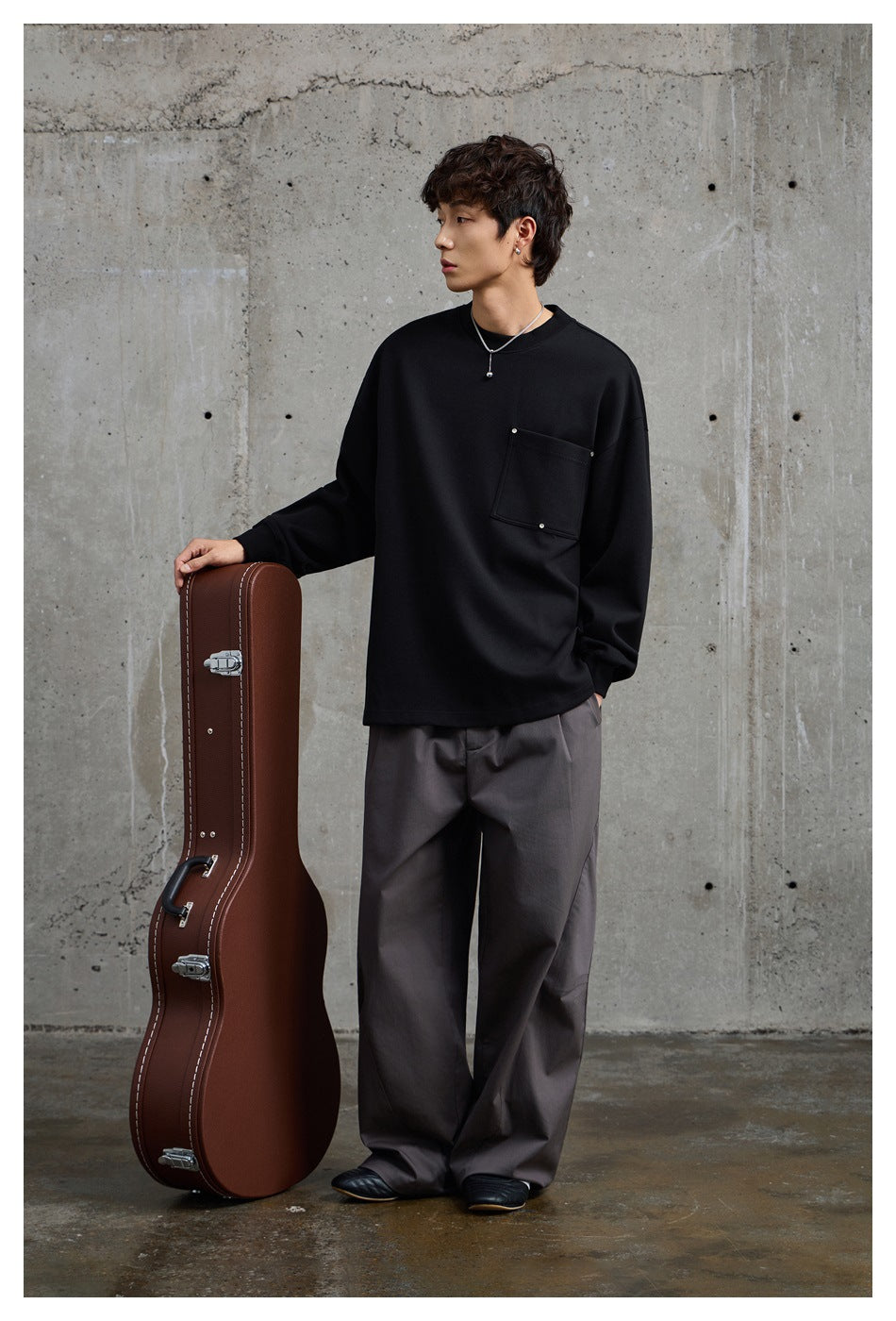 Full-length model wearing a black oversized crewneck sweatshirt with a square chest pocket and loose gray trousers.