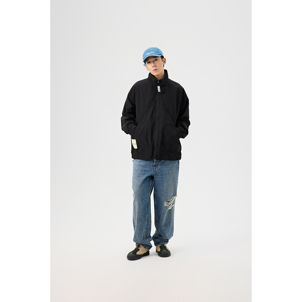Full-length view of a model wearing a black zip-front jacket with a high collar, blue jeans, and a blue cap.