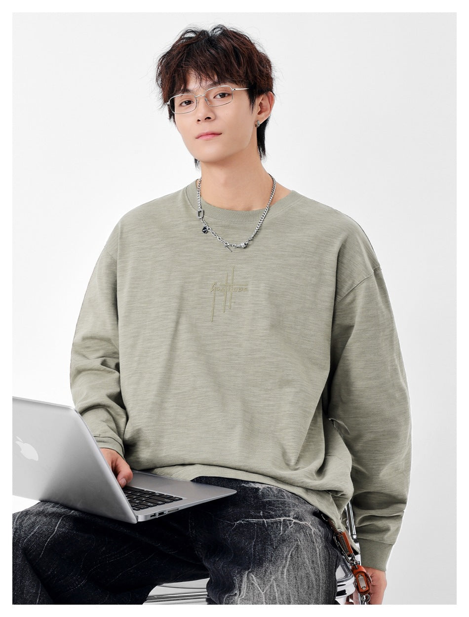 Model seated in a sage-gray oversized crewneck shirt with subtle chest embroidery and ribbed cuffs.