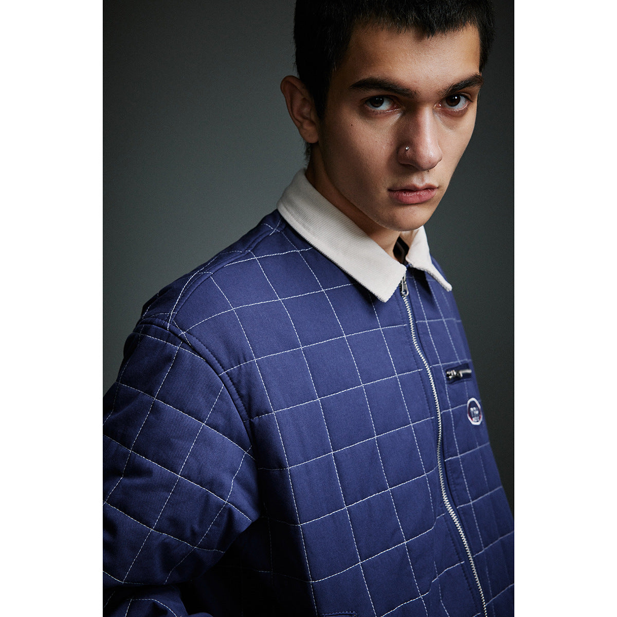 Close-up of a model wearing a navy quilted jacket with white grid stitching, a cream collar, front zipper and chest details.