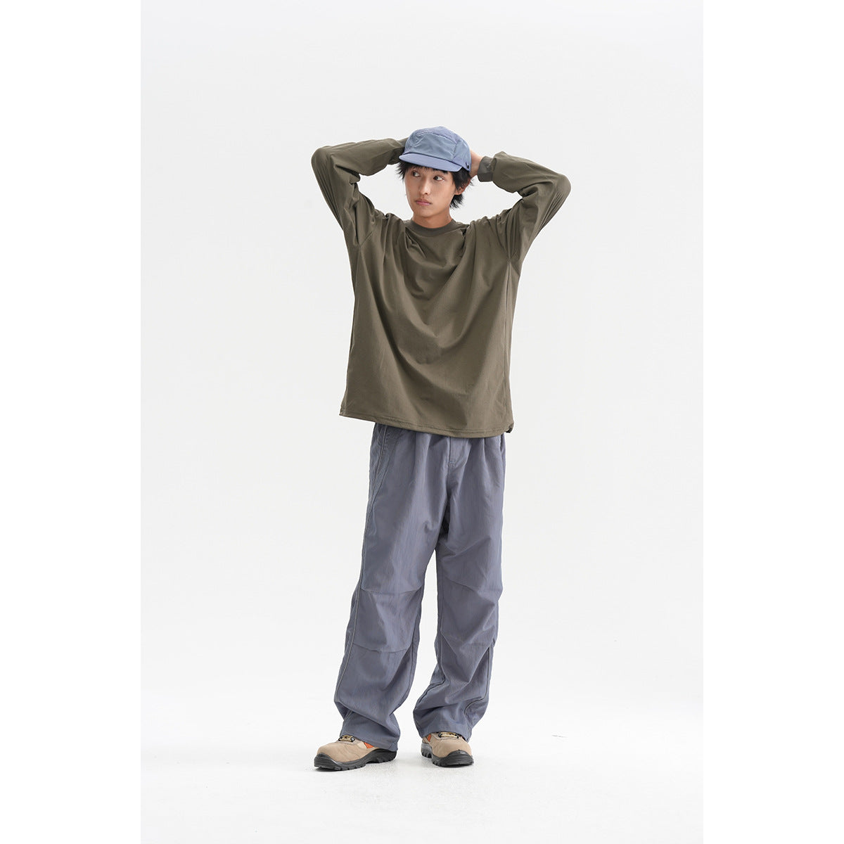 Full-length studio view of a model in an olive long-sleeve shirt, slate-blue loose pants, and a blue cap.