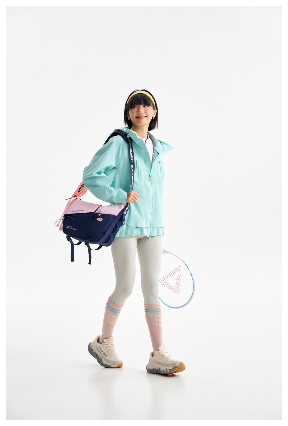 Model wearing an aqua hooded zip jacket with white leggings, carrying a sports bag and racket.