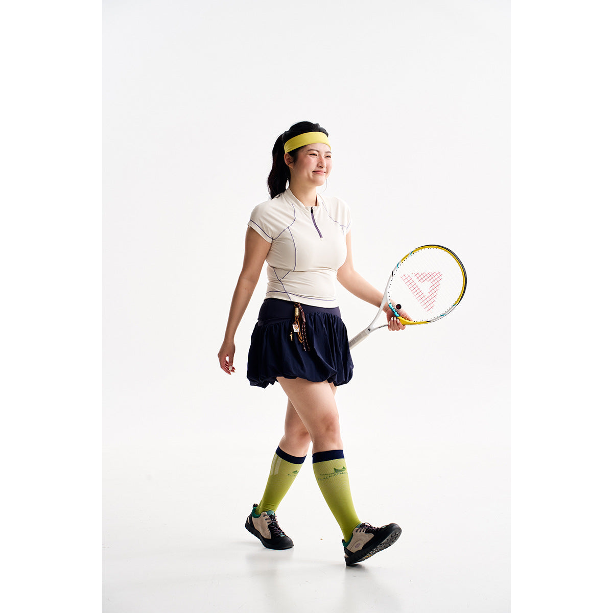 Model wearing a white zip-neck athletic top, navy skirt and yellow headband while holding a tennis racket.