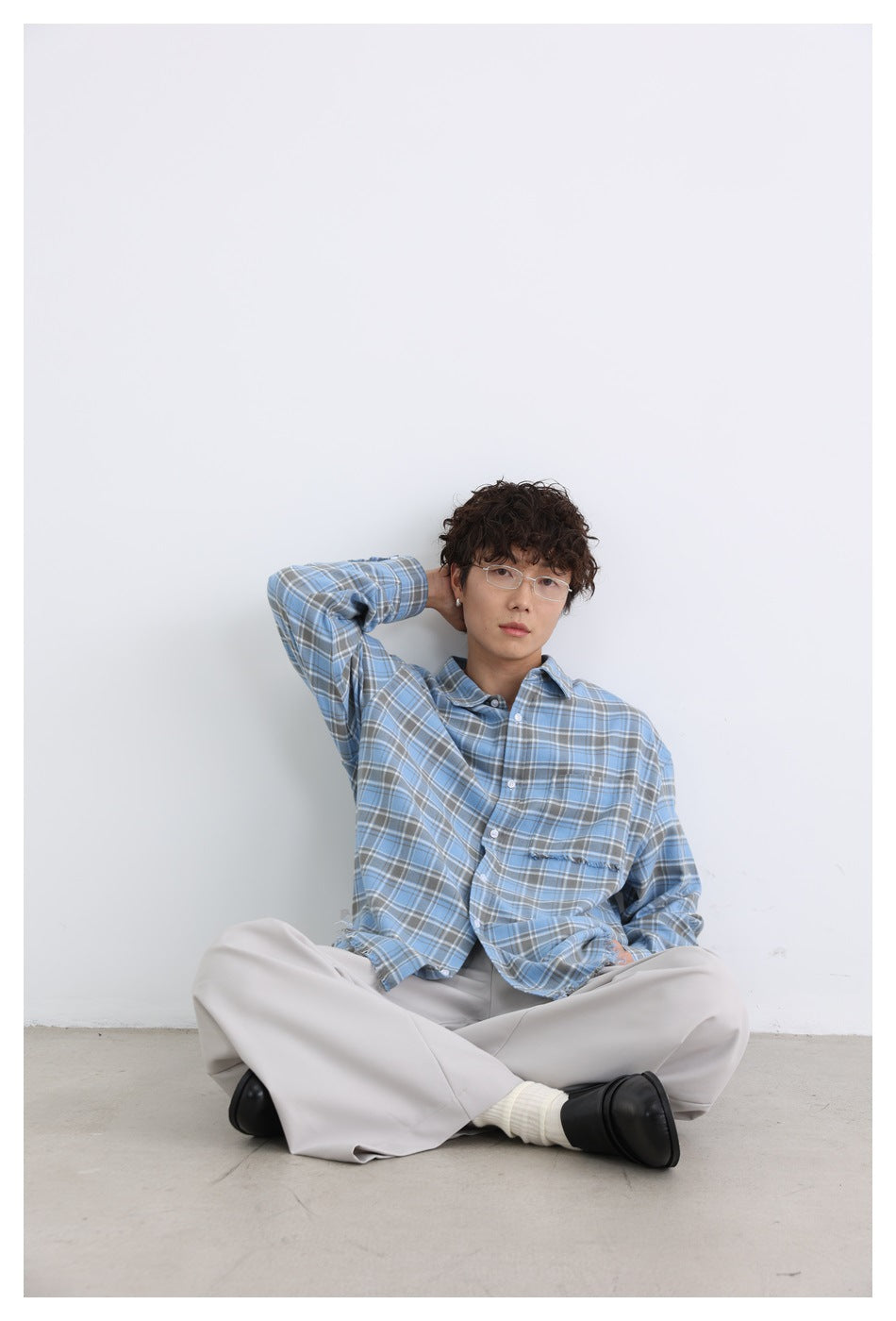 Model seated against a white wall wearing a blue and gray plaid button-up shirt with a chest pocket.