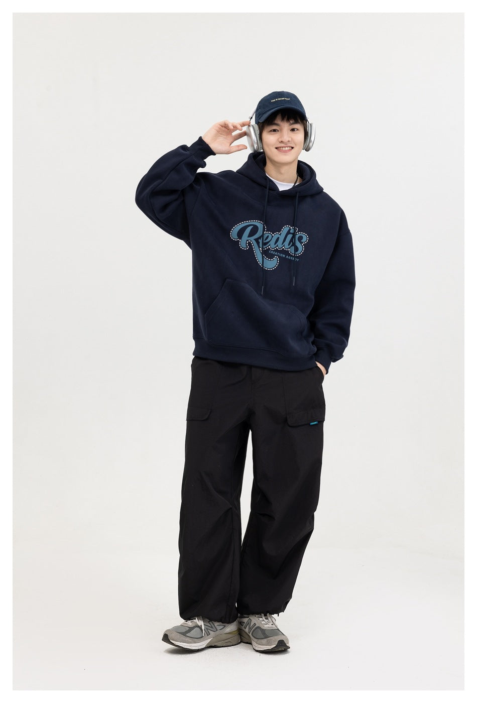 Smiling model in a navy oversized hoodie with blue script graphic, black cargo pants, cap and headphones