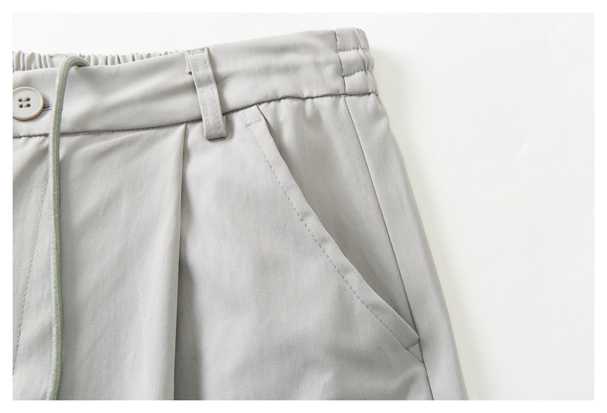 Close-up of light gray trousers showing the waistband, belt loop, button edge, drawstring and slanted pocket.