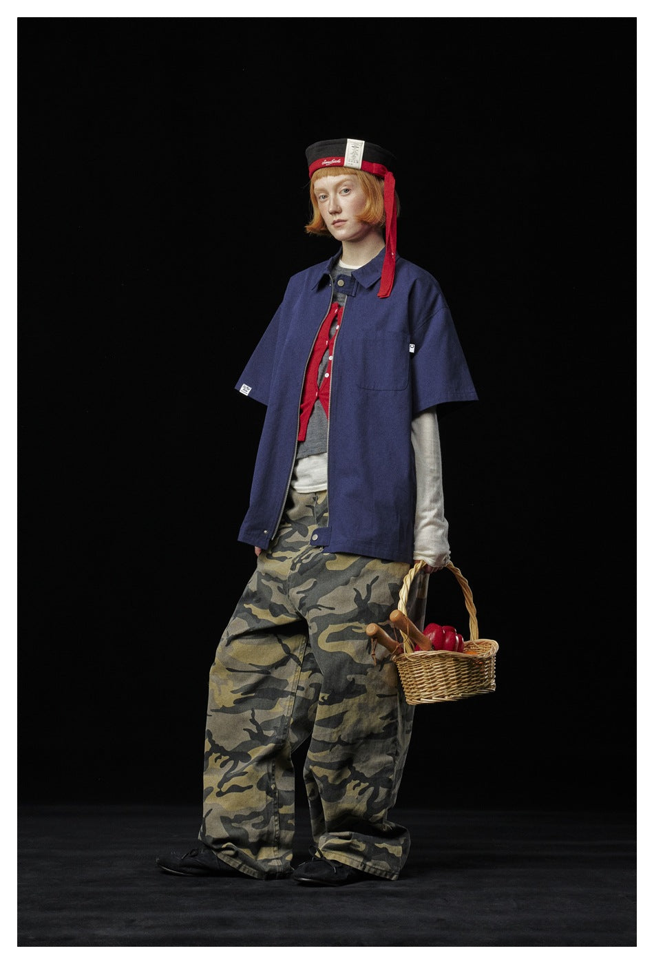 Model wearing a blue short-sleeve zip shirt open over layers with camouflage pants, angled view holding a basket