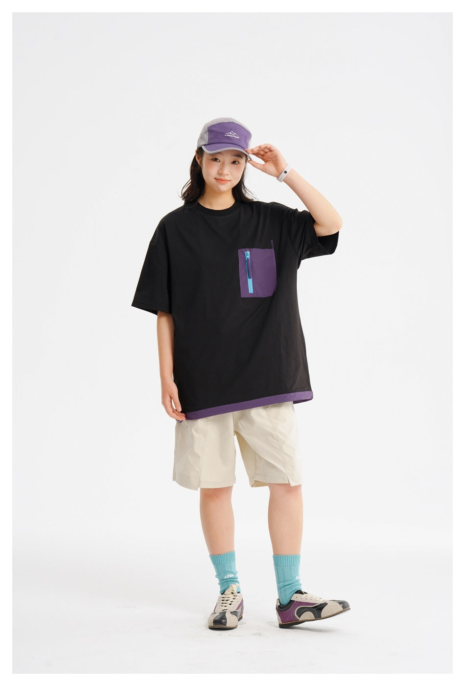 Person in a black T-shirt with a purple zip pocket and hem, hand on cap brim, with beige shorts and teal socks