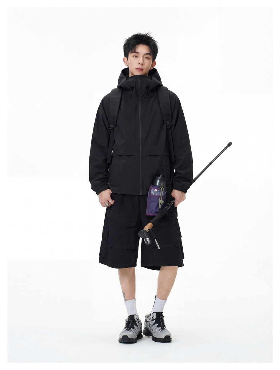 Full-length model in a black hooded jacket and cargo shorts, carrying a backpack and a trekking pole.