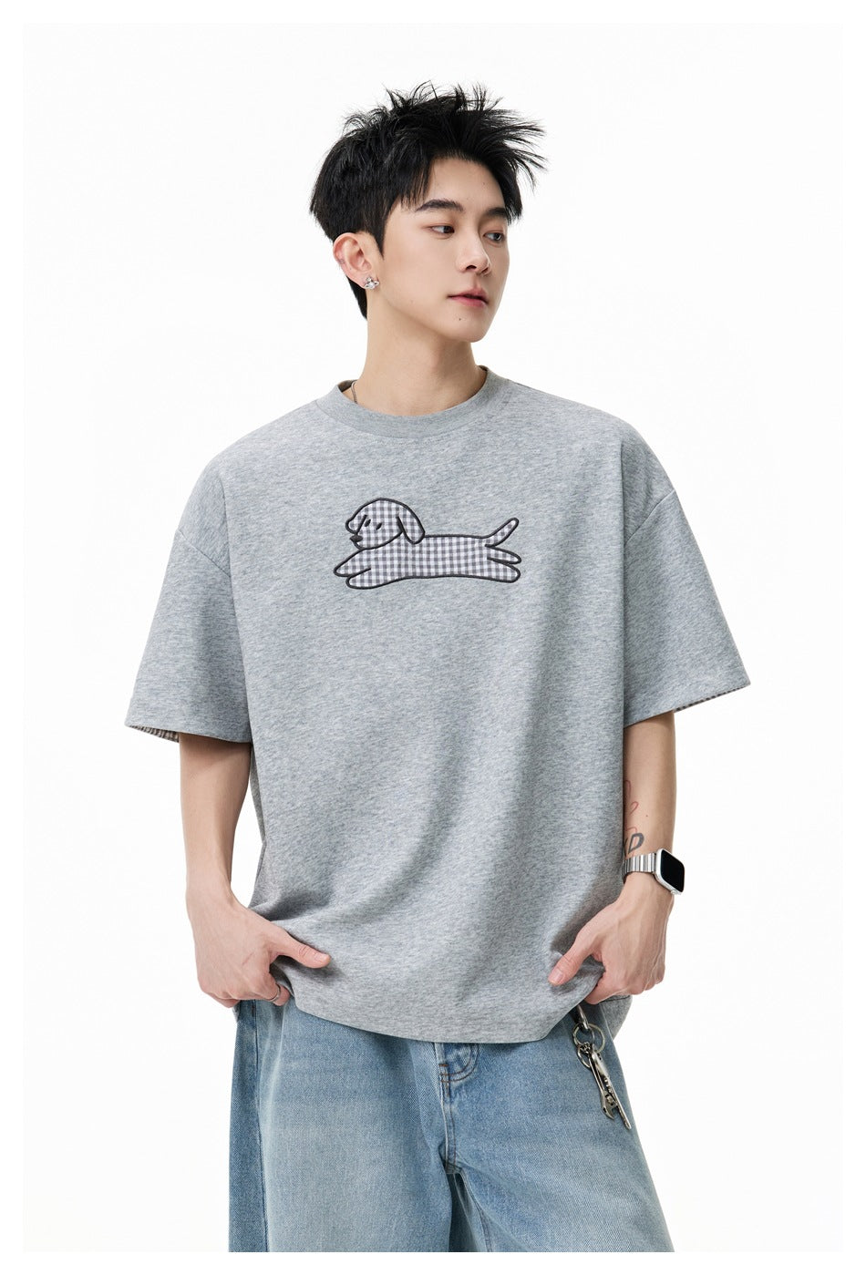 Model wearing an oversized heather-gray T-shirt with a checked dog graphic on the chest and blue jeans.