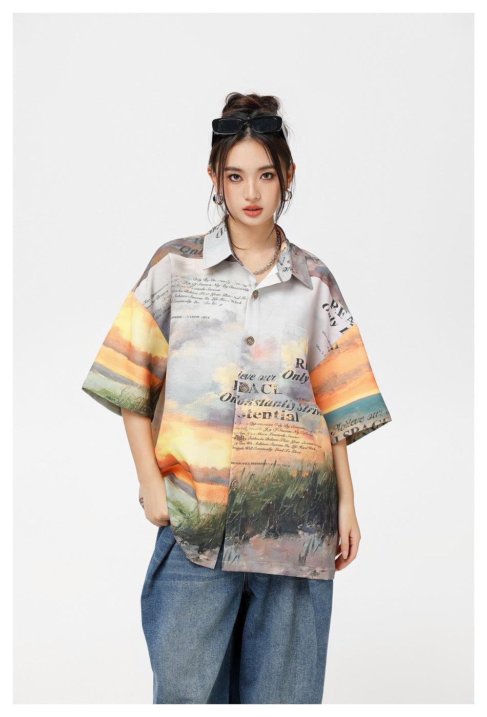 Model wearing an oversized short-sleeve shirt with a painterly sunset landscape and black text print, paired with blue jeans.