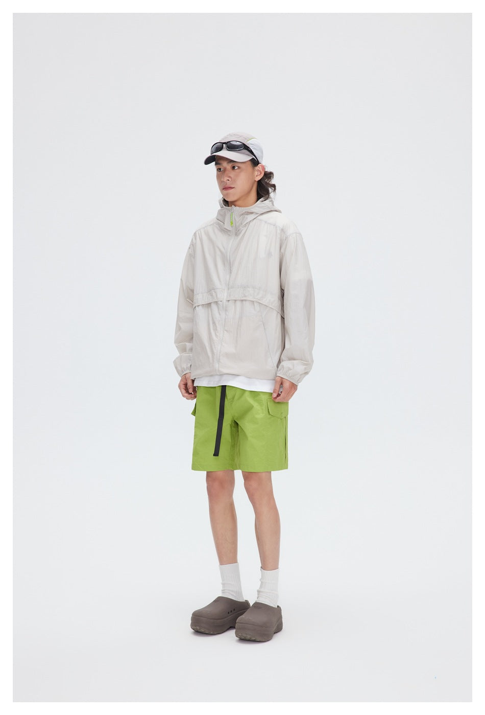 Model wearing a light beige hooded zip jacket with green shorts against a pale background.