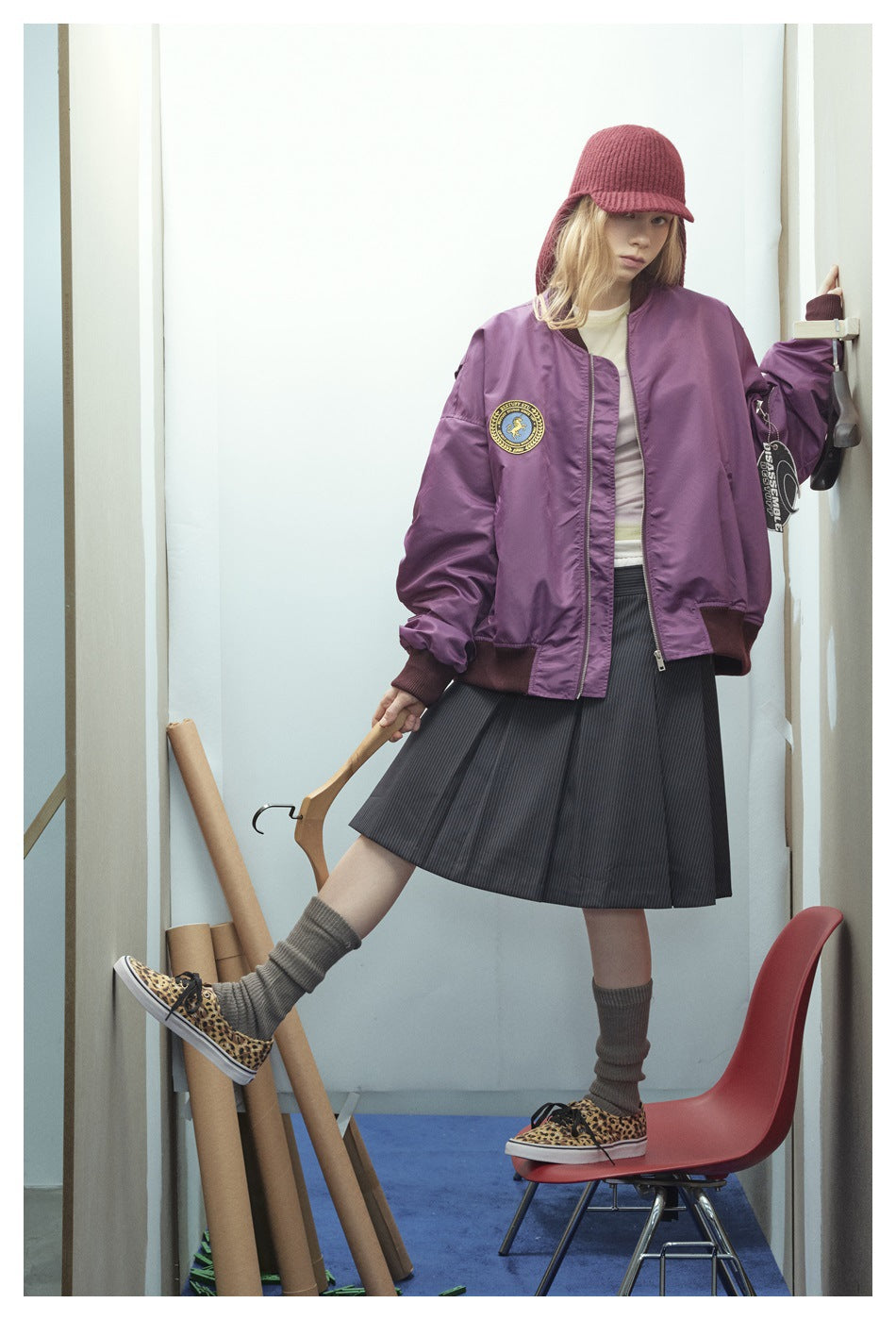 Model wearing a purple bomber jacket with a round chest patch, zip front, dark ribbed trim, and a hanging sleeve tag.