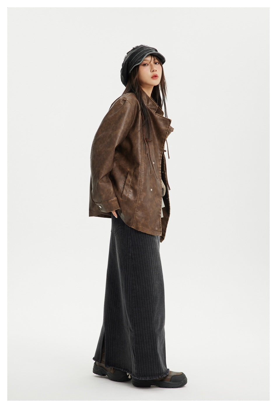 Side view of model in a brown faux-leather-look jacket with long gray ribbed skirt, cap and chunky boots