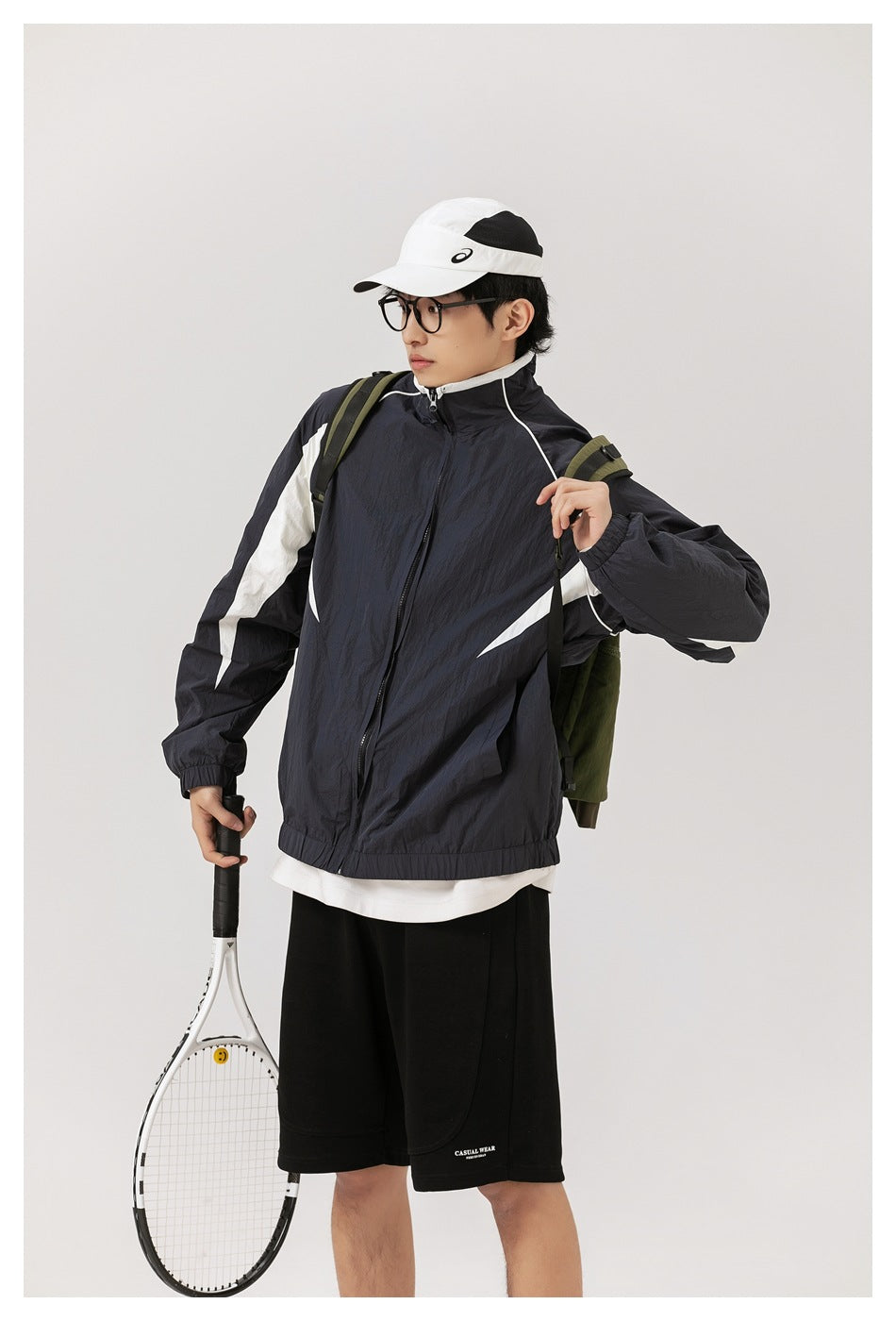 Model wearing a dark navy and white zip jacket with black shorts, carrying a backpack and holding a tennis racket.