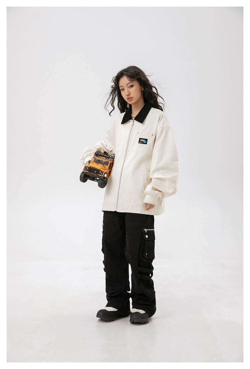 Model wearing an ivory zip-front jacket with a black collar and black cargo pants, holding a toy truck.