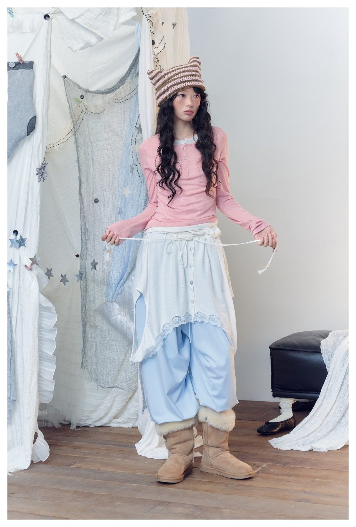 Model in pink henley top holding a skirt drawstring, with striped knit hat, light blue pants and tan boots
