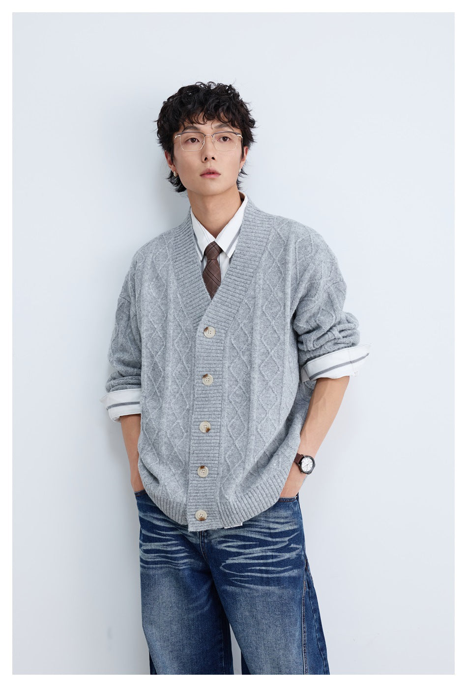 Man wearing a light gray diamond cable-knit button cardigan over a white shirt and brown tie with blue jeans