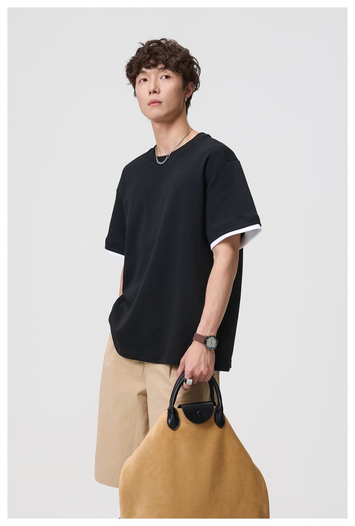 Model in black crewneck T-shirt with white layered sleeve hem, angled view with khaki shorts and tan bag