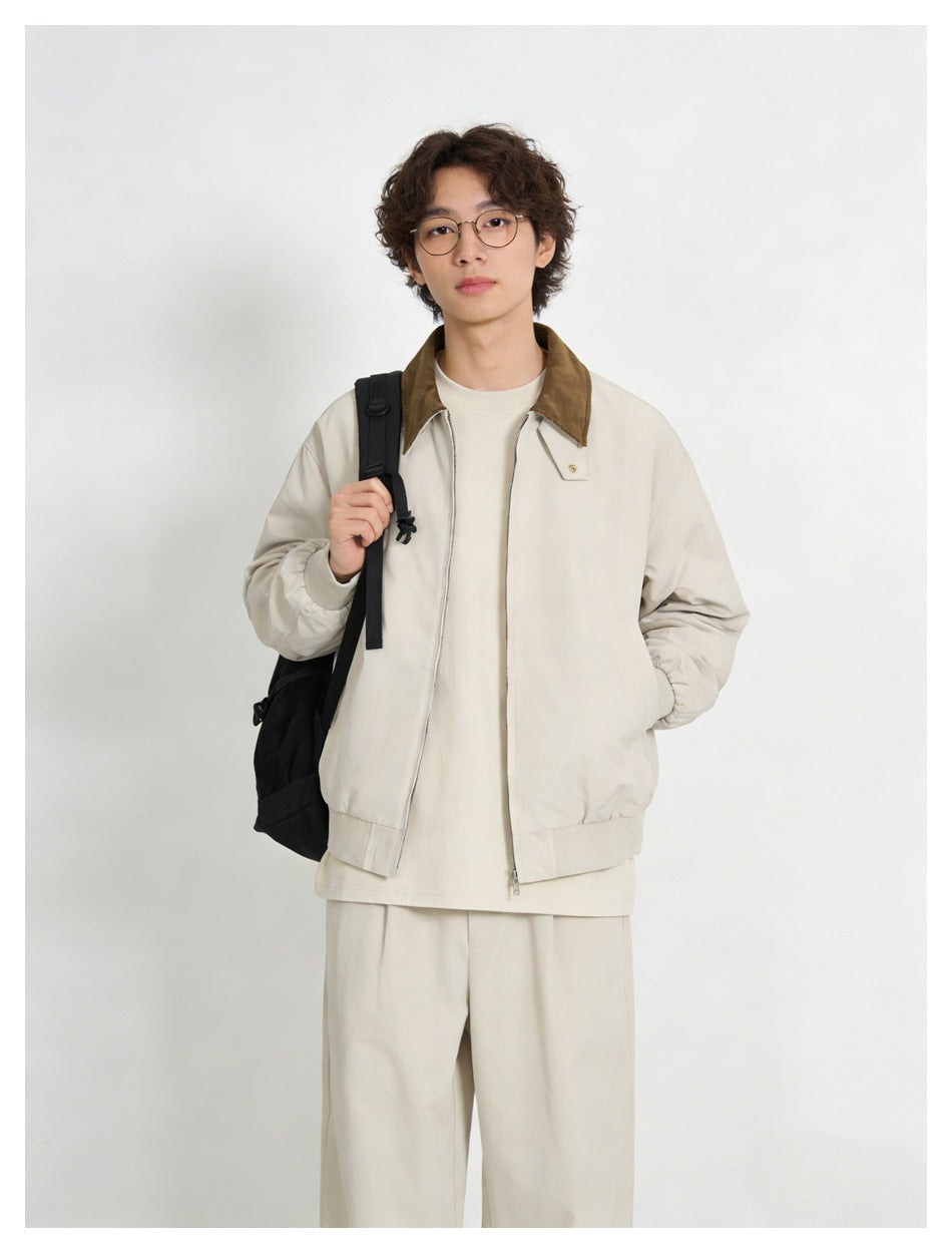 Man wearing beige zip jacket with brown collar over a cream tee, holding a black backpack strap, with beige trousers