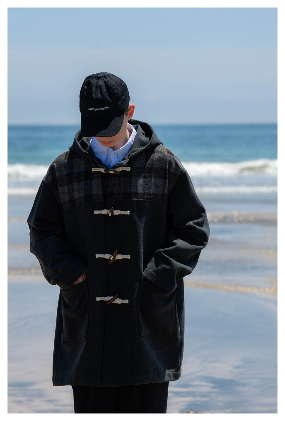 Close view of a person with a black cap wearing a charcoal toggle coat with plaid yoke and hands in the front pockets