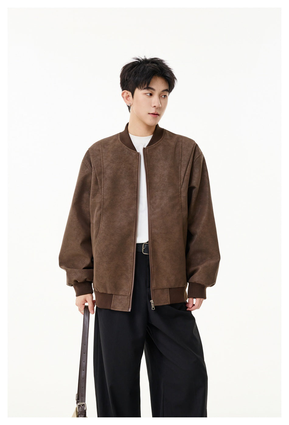 Man wearing a brown suede-look zip bomber jacket over a white tee and black wide-leg pants, holding a bag strap