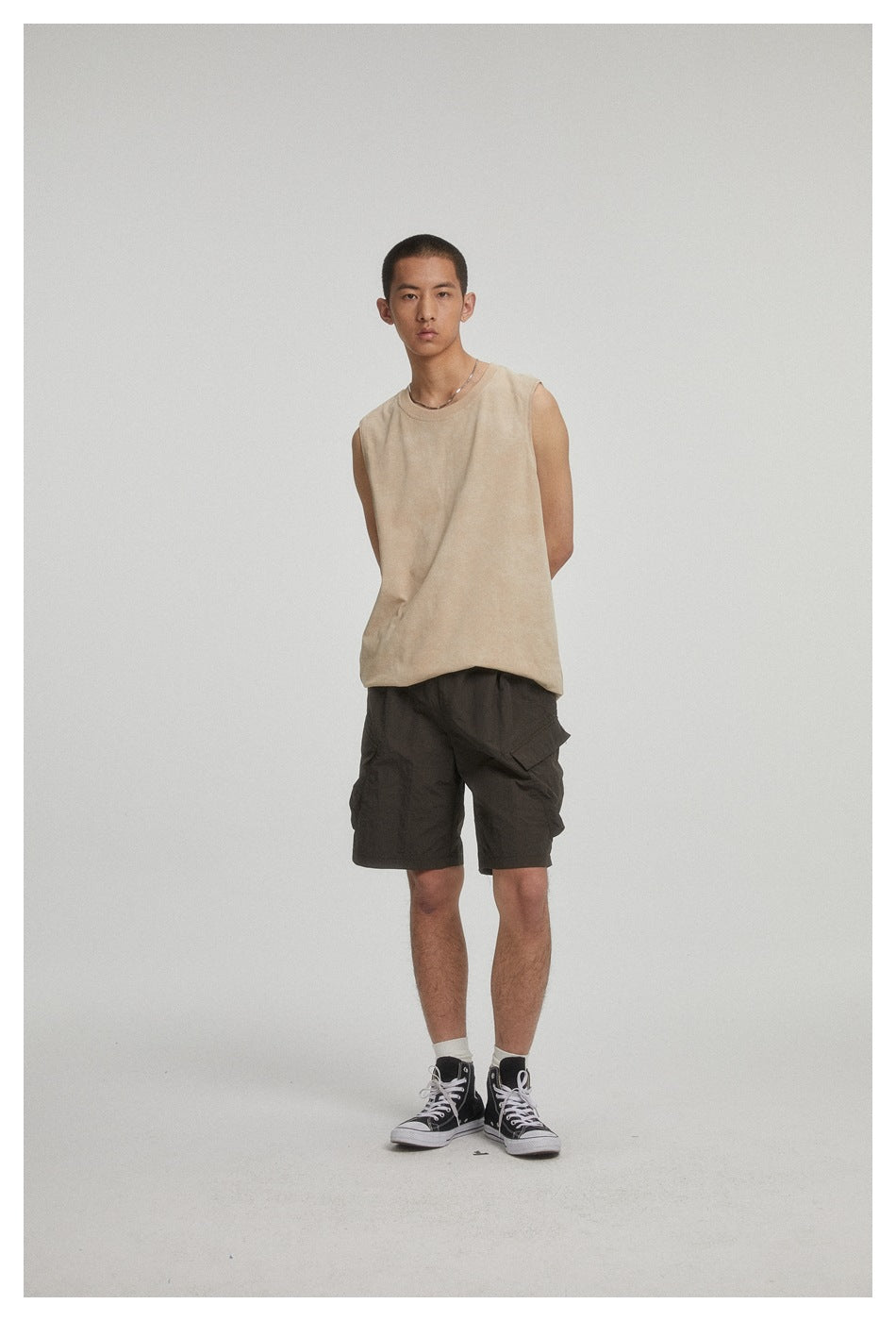 Full-length model wearing a beige sleeveless crew-neck shirt, dark cargo shorts, and black high-top sneakers.