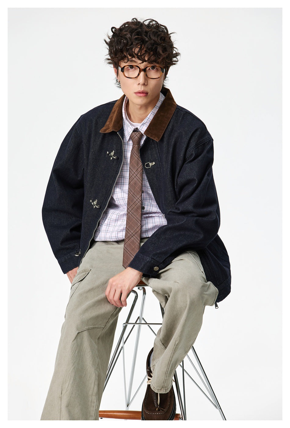 Man seated on a stool wearing a navy denim jacket with brown collar over a plaid shirt and tie