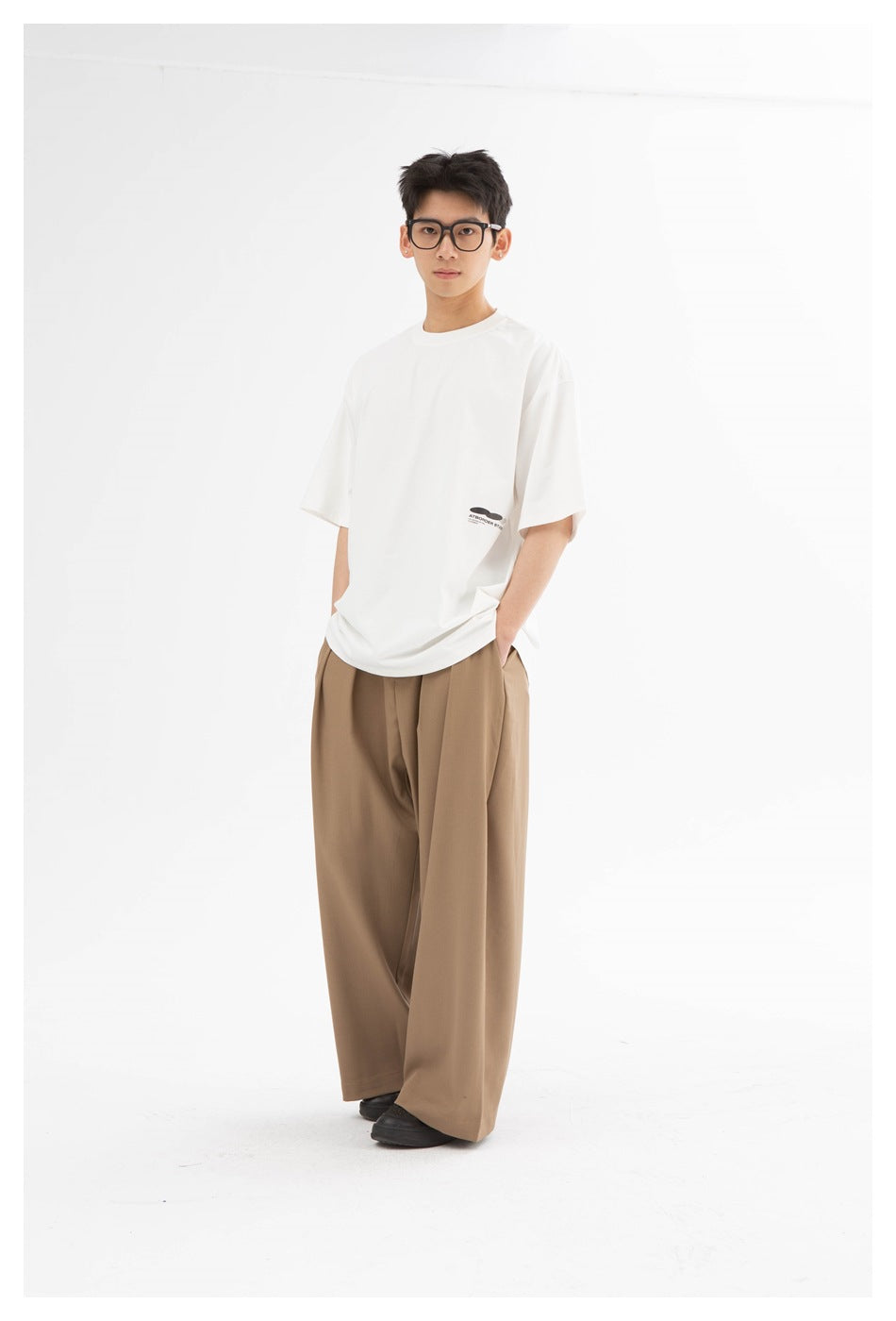 Model standing in a white oversized crew-neck t-shirt with lower-hem print, wide-leg tan trousers and black shoes