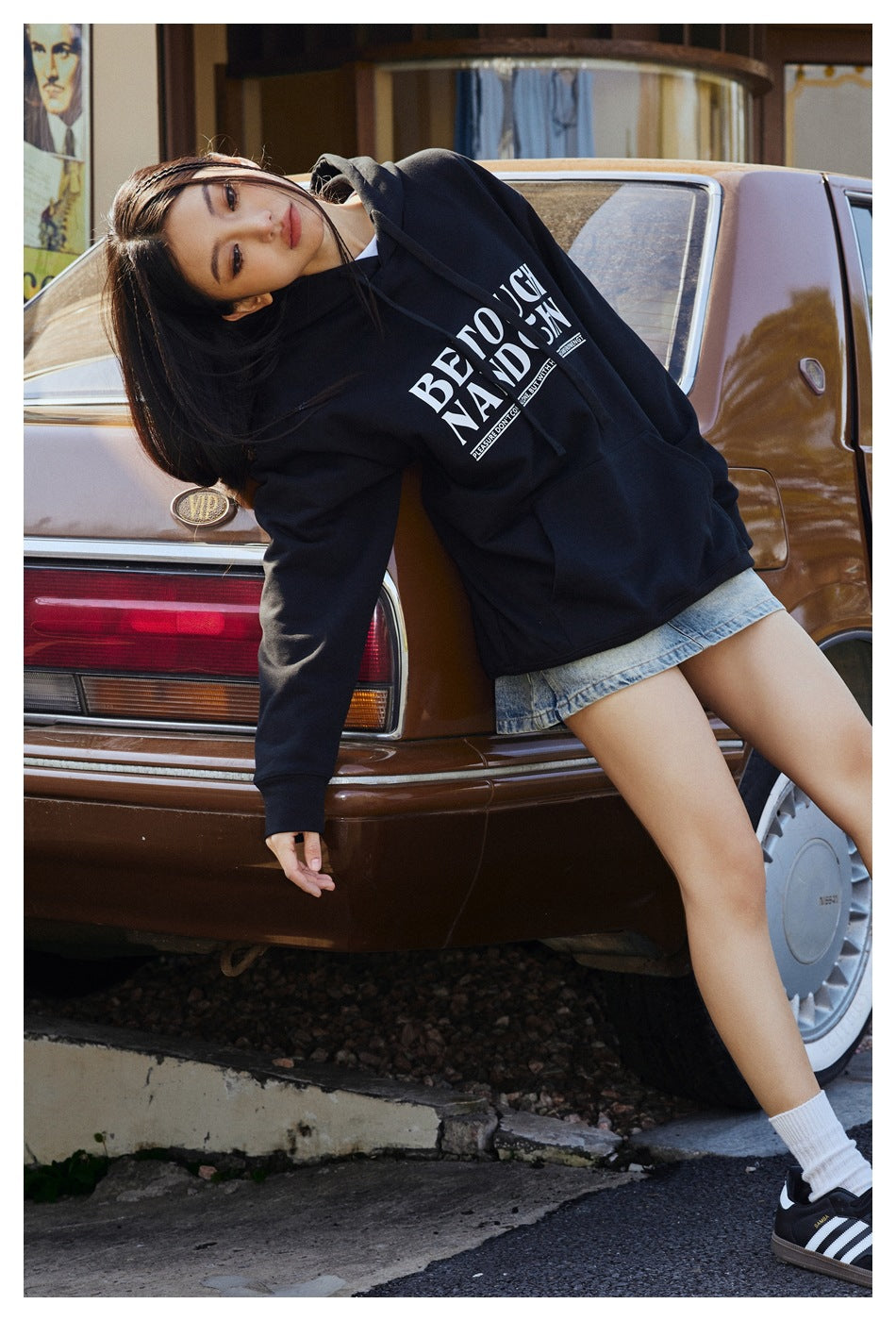 Model reclines against a vintage car wearing a black oversized graphic hoodie and denim shorts.
