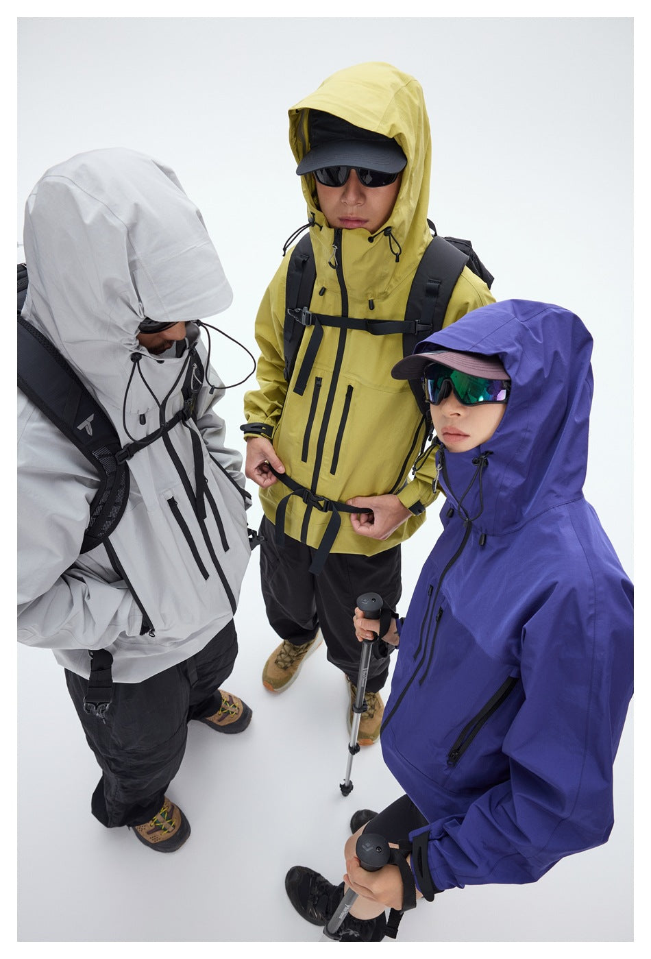 Three hikers wear hooded technical jackets in white, yellow, and purple, with dark pants, sunglasses, and trekking gear.