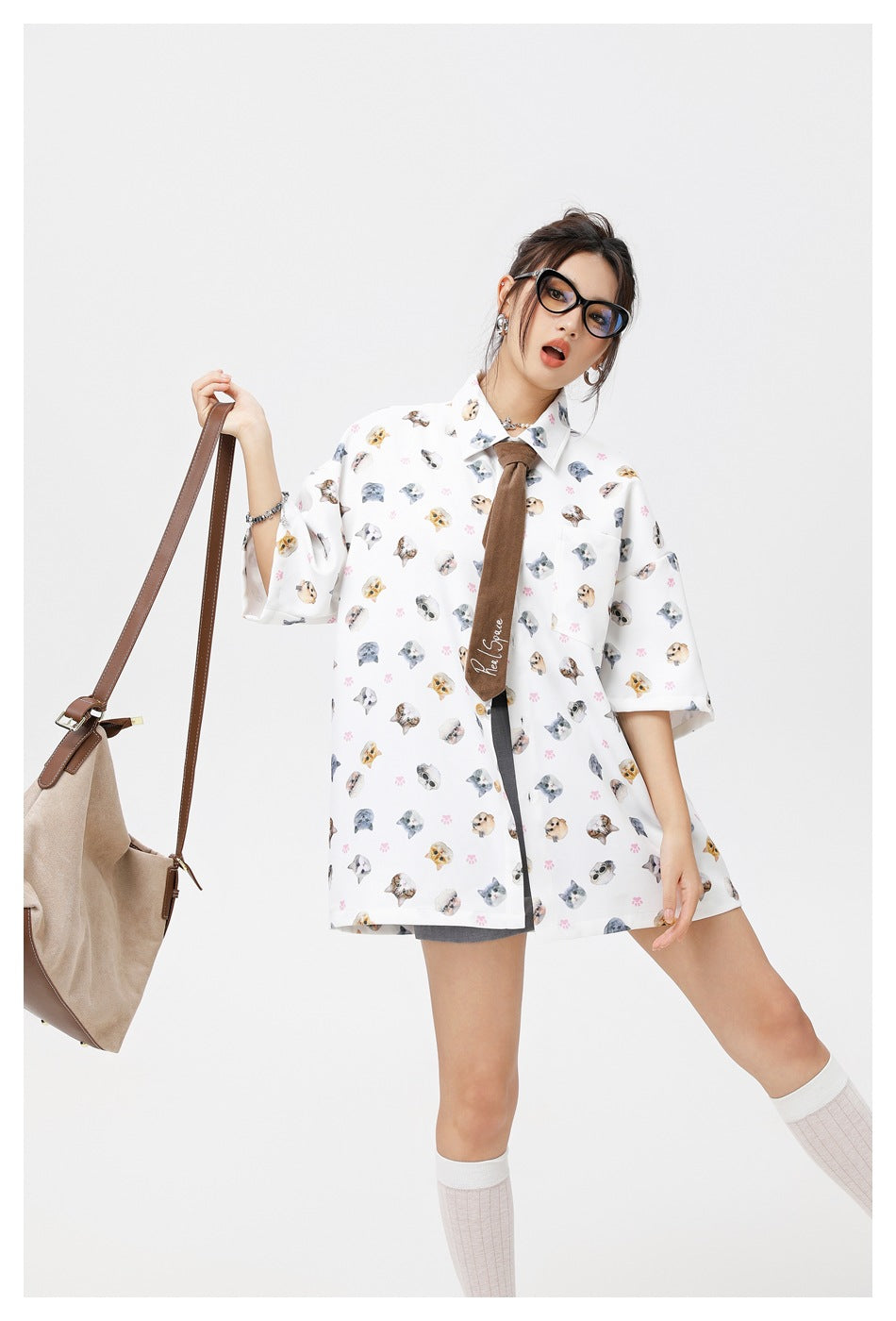 Model wearing a white oversized short-sleeve shirt covered in cat faces and pink paw prints, with a brown tie.