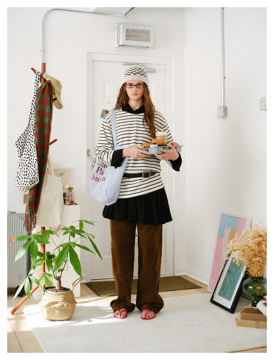 Full-length model wearing a loose cream top with thin black stripes, black collar and cuffs, and brown trousers.