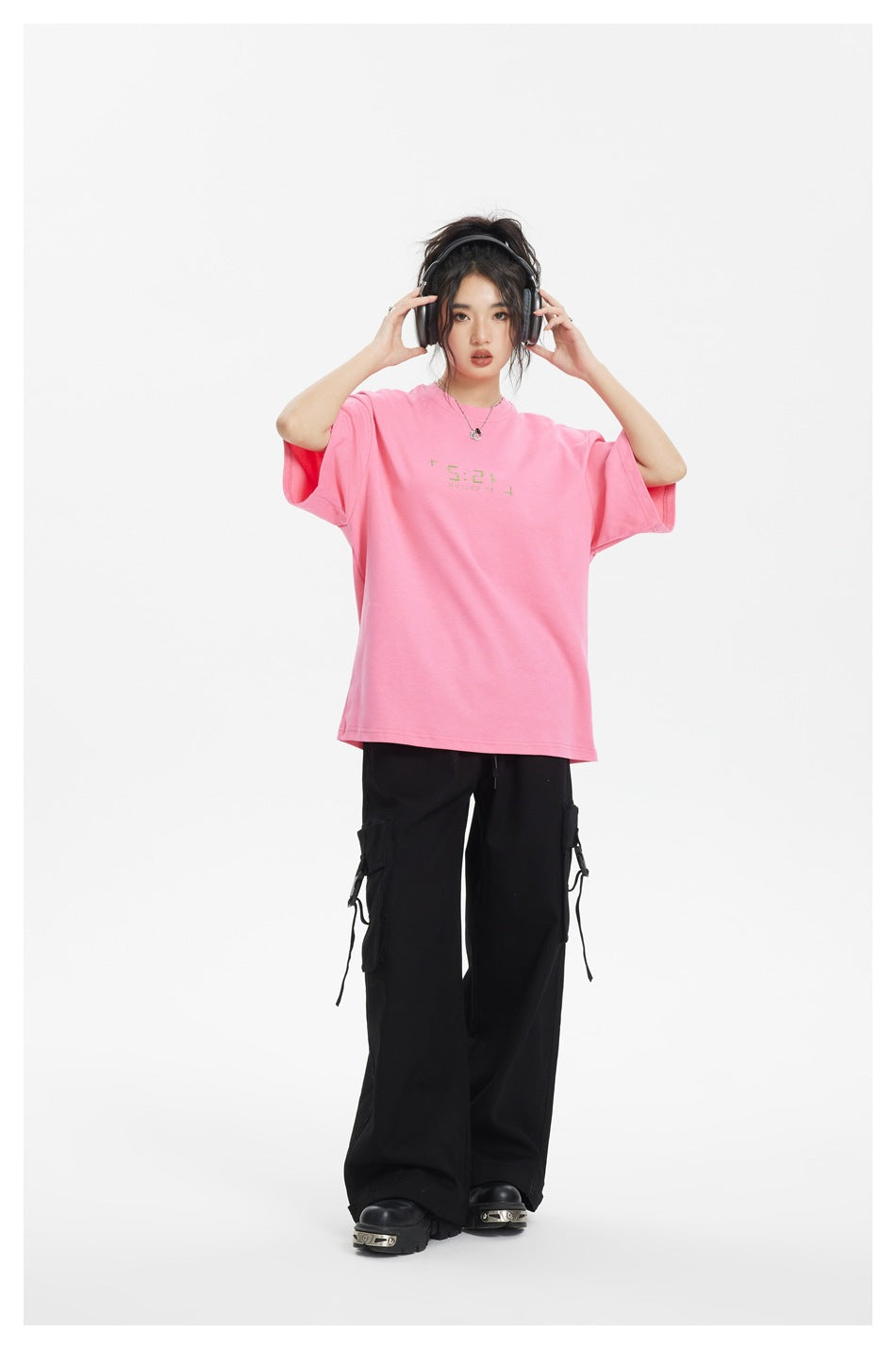 Model in an oversized pink graphic T-shirt with black cargo pants, hands on headphones