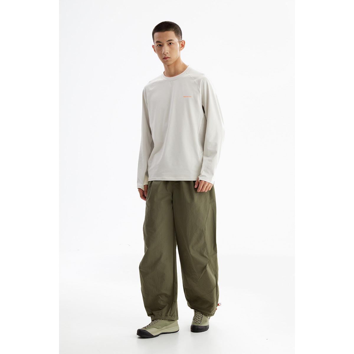 Model wearing an off-white long-sleeve top with a small orange chest mark and loose olive trousers.