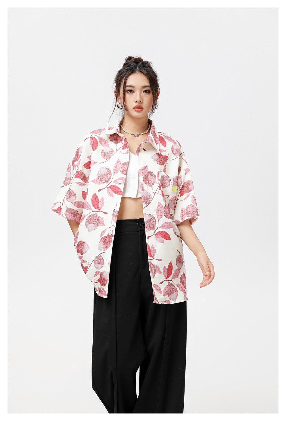 Model wearing an open white short-sleeve shirt with pink leaf print over a white crop top and black trousers.