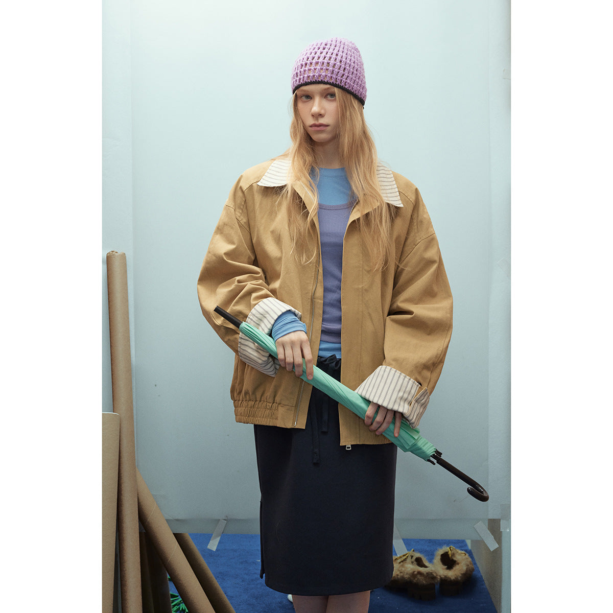 Model wearing a tan jacket with striped collar and cuffs over blue layers, paired with a dark skirt and pink knit beanie.