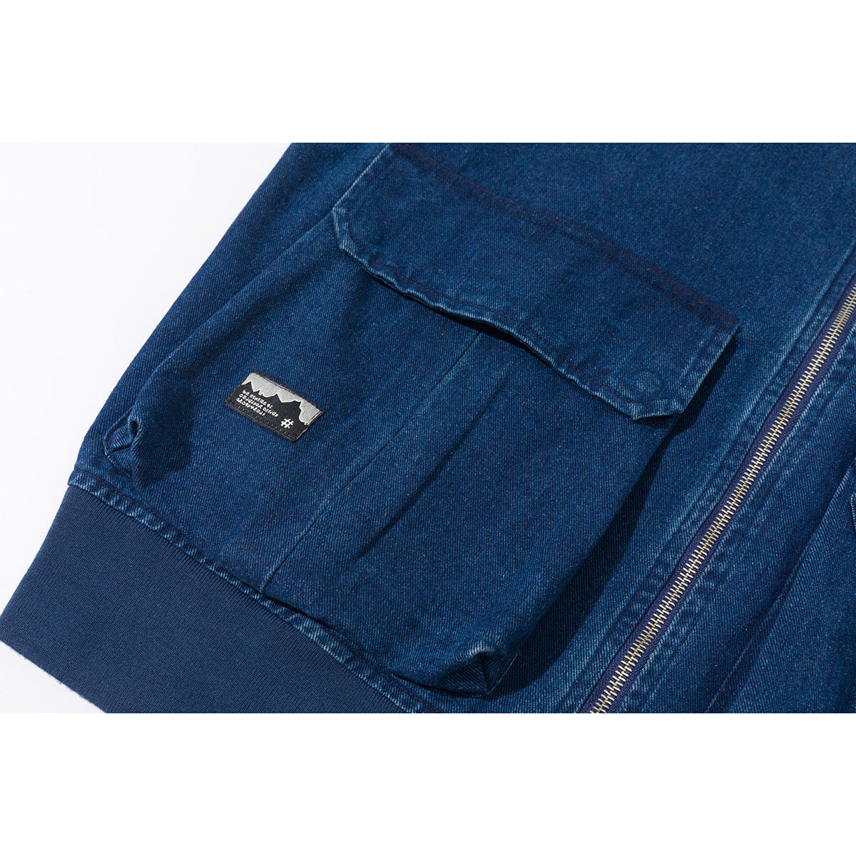 Close-up of a blue denim jacket's flap pocket, small patch, ribbed cuff, and metal zipper.