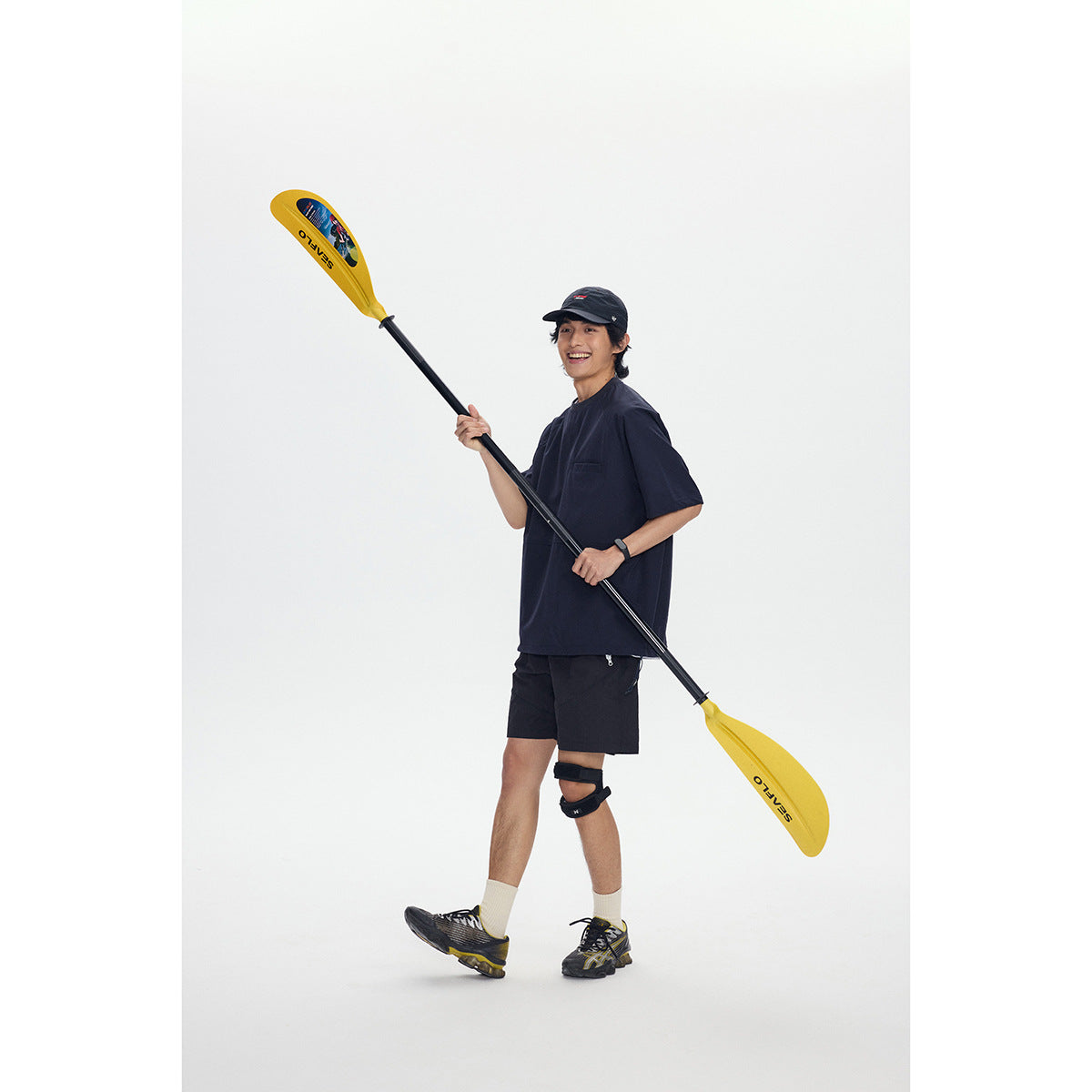 Model wearing a loose navy short-sleeve shirt and black shorts, holding a yellow paddle against a white background.