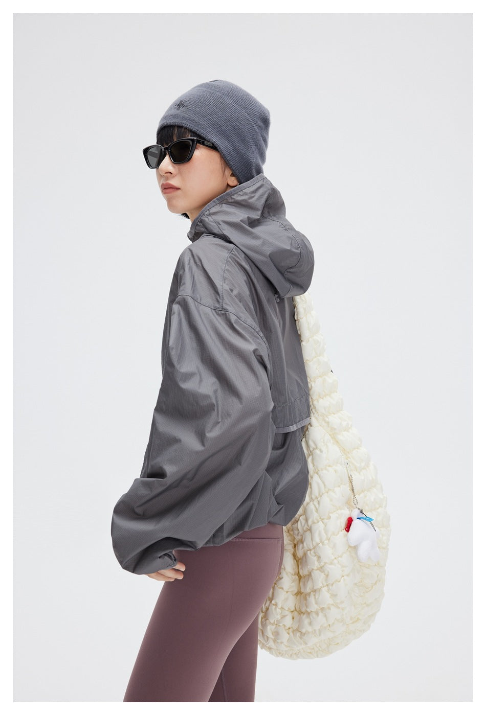 Three-quarter rear view of a model wearing a gray hooded windbreaker and mauve leggings, with a white shoulder bag.