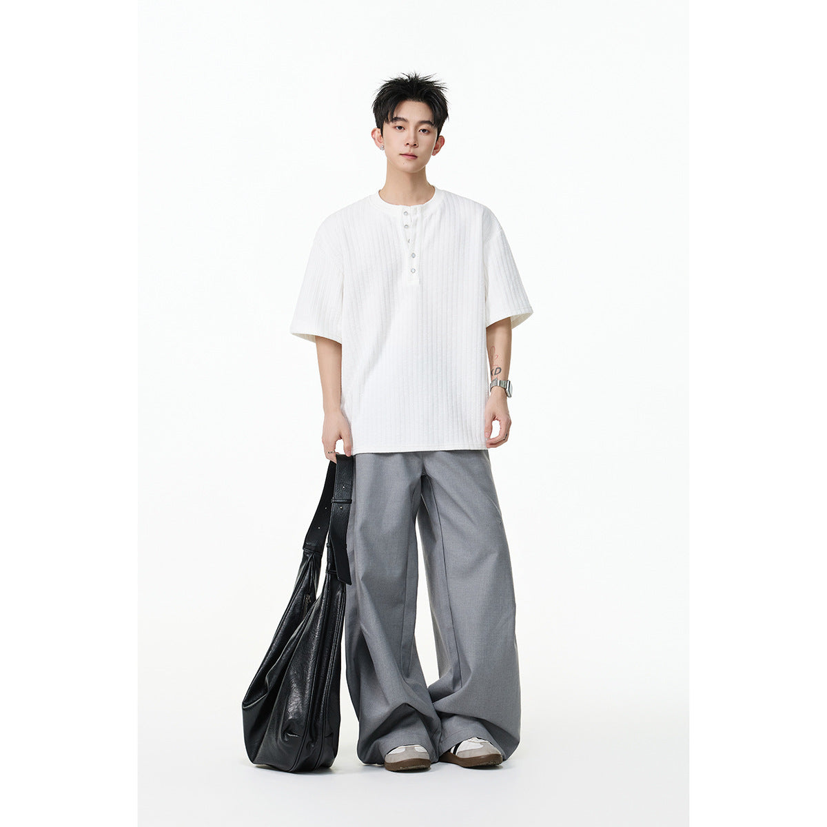Model wearing a loose white ribbed short-sleeve henley with gray wide-leg trousers, holding a black bag.