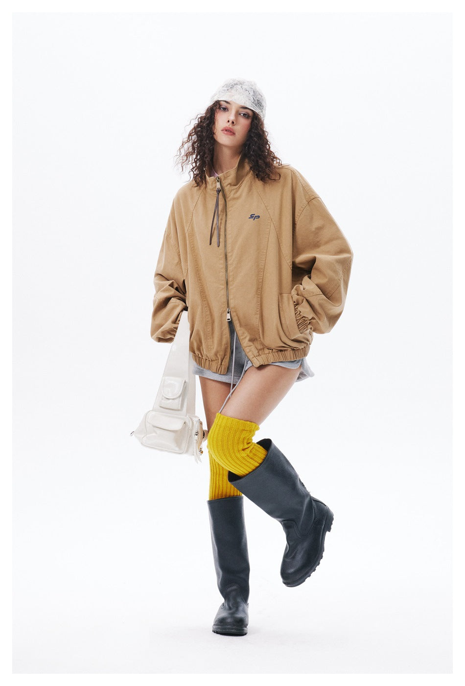 Model wearing an oversized tan zip-front jacket with a stand collar and elastic hem, styled with gray shorts and black boots.