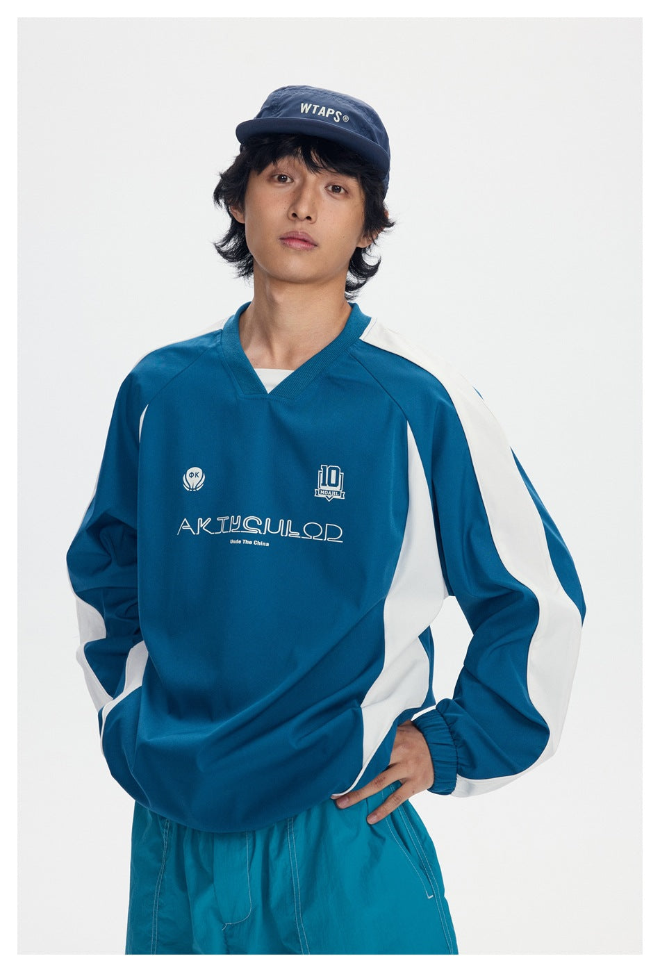 Model wearing a teal V-neck pullover with white sleeve and side panels, chest graphics, and a navy cap.