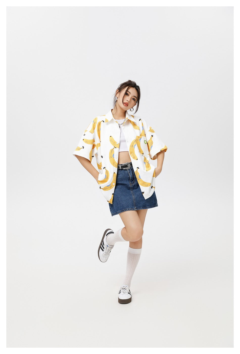 Model posing in a white banana-print short-sleeve shirt and denim mini skirt against a light background.