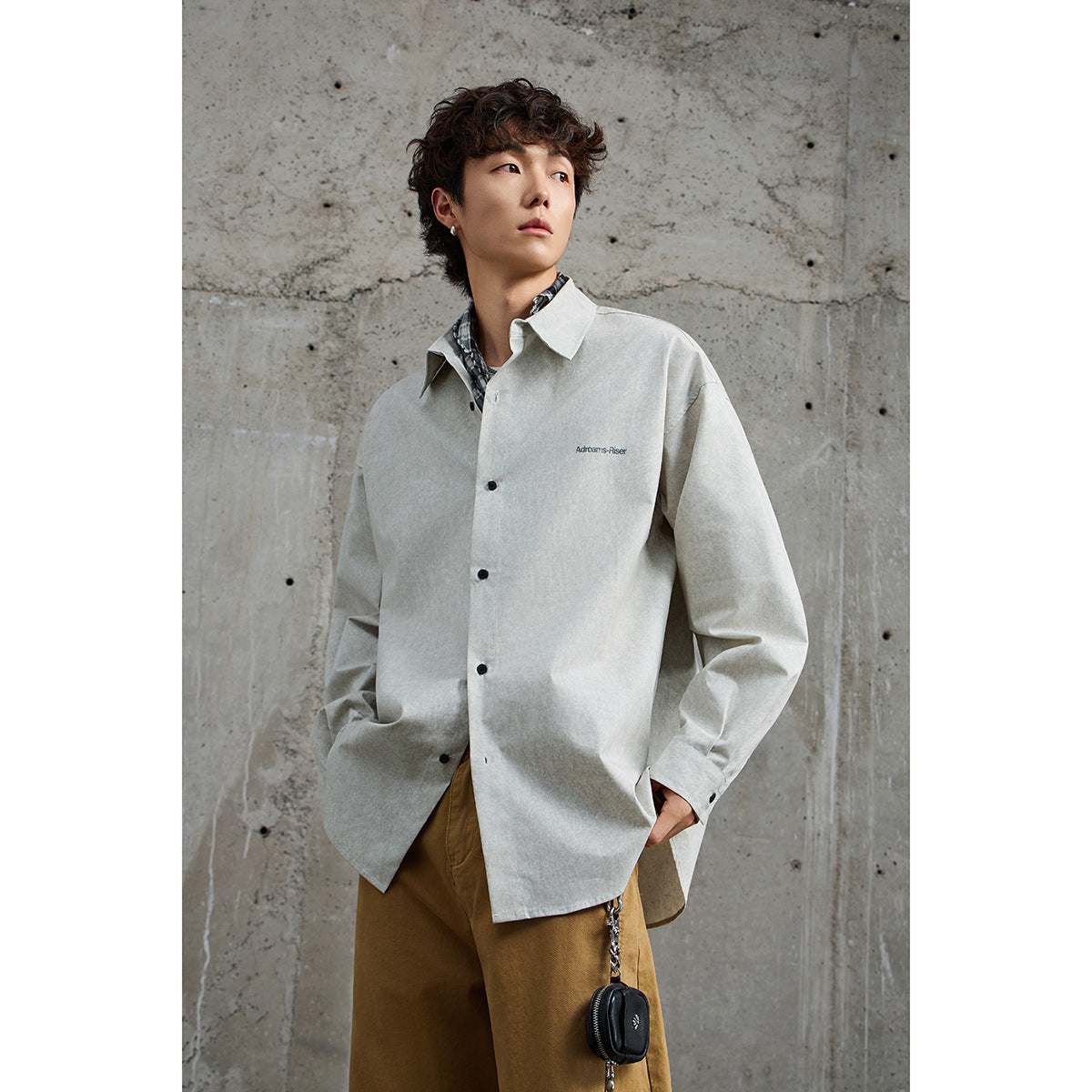Model wearing a pale gray button-up shirt with small chest lettering and tan trousers, shown from the front.