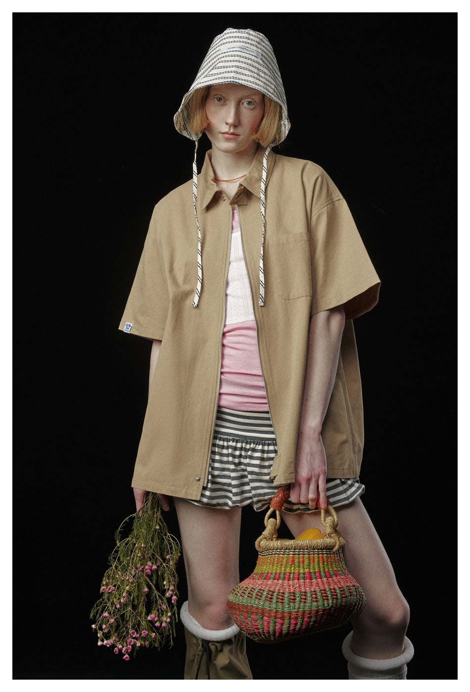 Model wearing an oversized tan short-sleeve zip jacket, striped bucket hat and shorts against a black background.