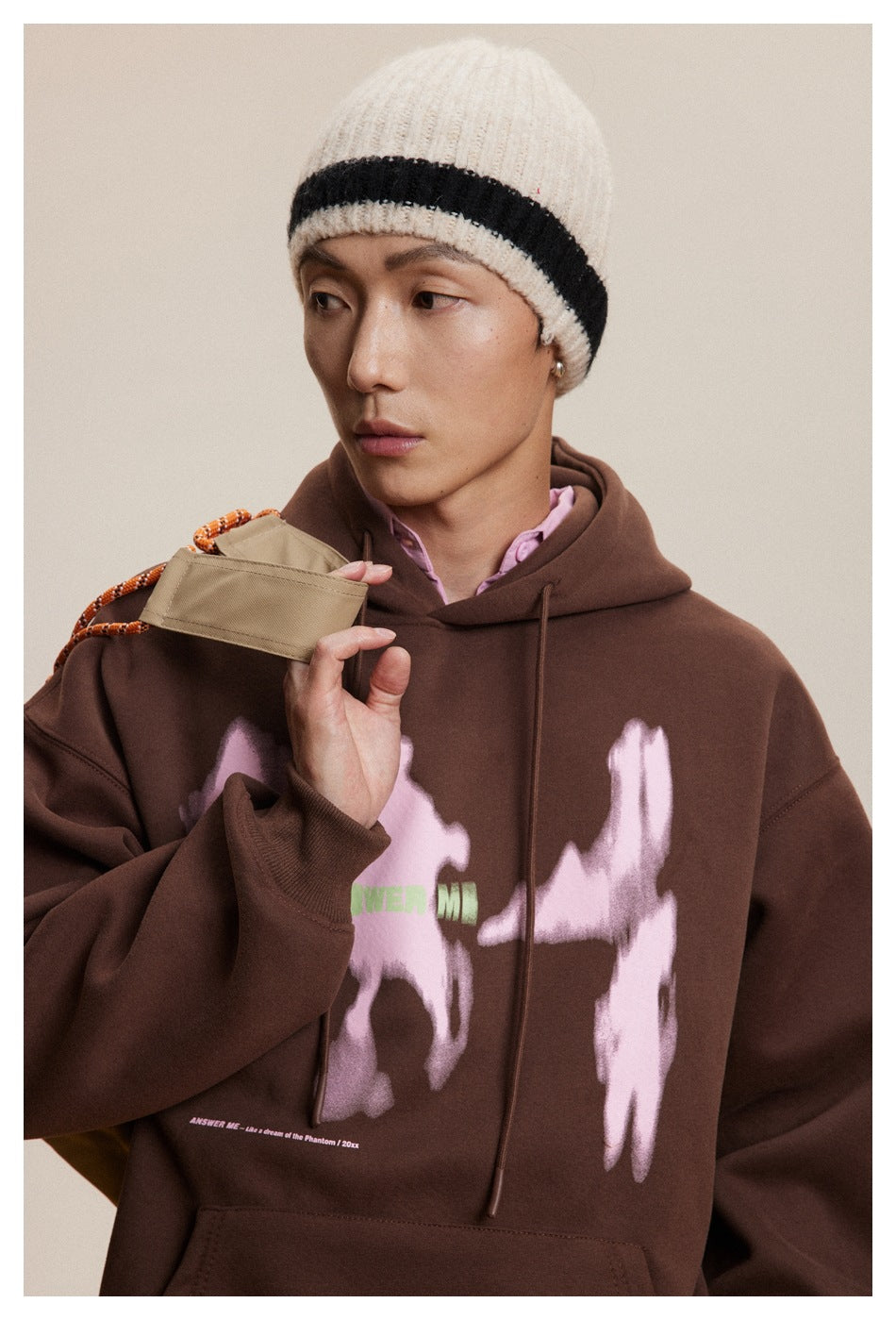Close-up of model in a brown hoodie with pink blurred print and green lettering, wearing a striped beanie