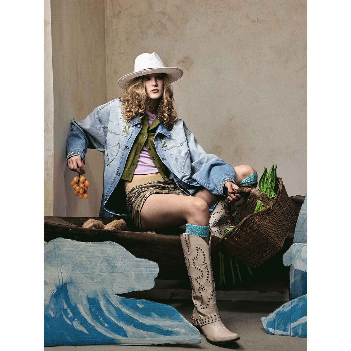 Model seated in an oversized light blue embroidered denim jacket, striped shorts, white hat and tall beige boots.